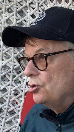 Foto von Ulrich Mill. Er sitzt in einem großen Korbstuhl, blickt an der Kamera vorbei, trägt eine Cap und eine braune Brille. Seine Lippen sind so geformt, als erzähle er gerade etwas. 