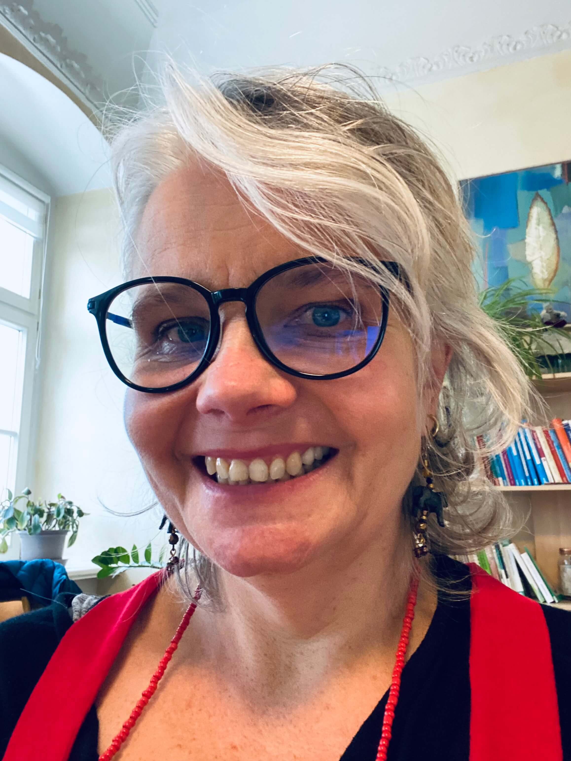 Selfie von Vera Nahrath. Sie lächelt in die Kamera und trägt eine große, schwarze Brille und eine rote Perlenkette. Hinter ihr sind ein buntes Büchergestell und Zimmerpflanzen.