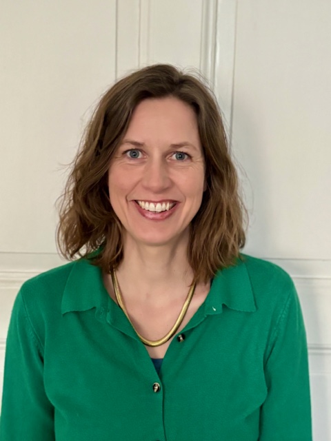 Foto von Julia Reineke. Sie steht vor einer weißen Wand, lächelt in die Kamera, trägt eine grüne Bluse und eine goldene Kette und ihre braunen, schulterlangen Haare offen.