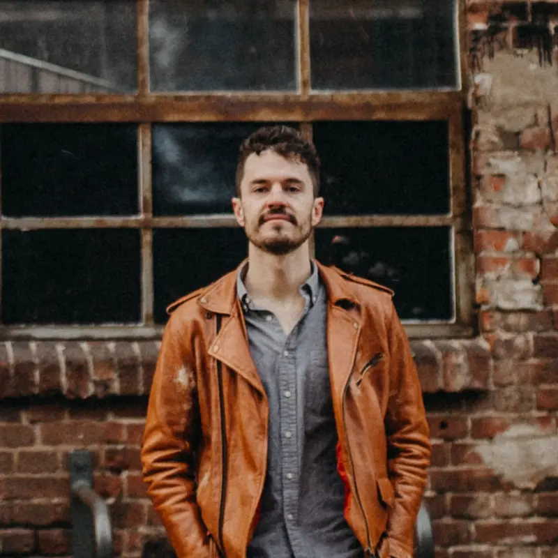 Ein Mann in grauem Hemd und brauner Lederjacke vor einem Backsteingebäude