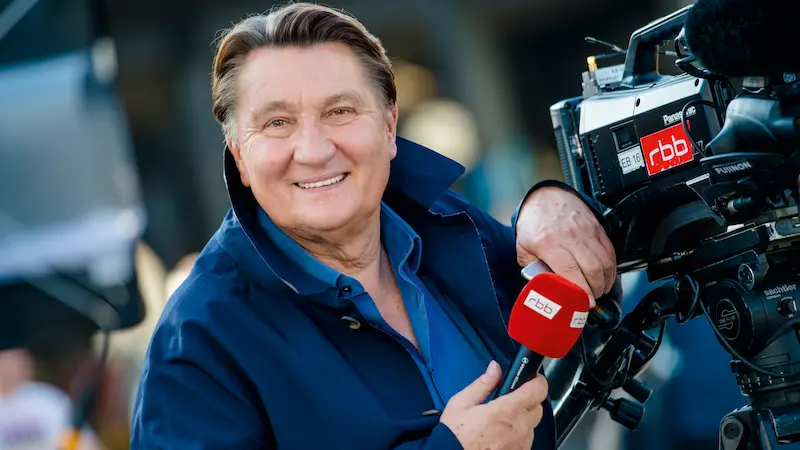  Ulli Zelle mit rbb-Mikro in der Hand, lehnt sich an eine rbb-Kamera