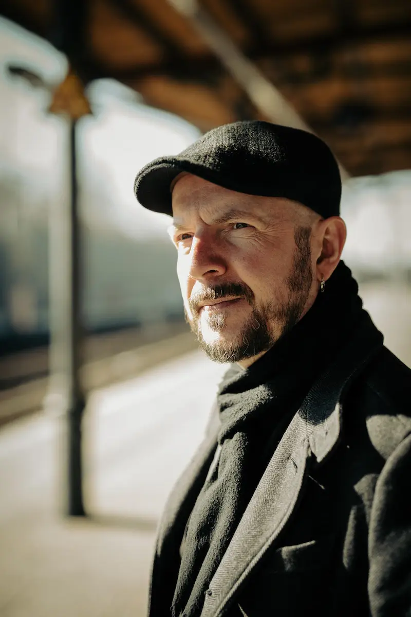 Porträtfoto von Marcel Krueger, er steht seitlich zur Kamera und schaut an ihr vorbei, er trägt eine schwarze Mütze, einen schwarzen Mantel und einen schwarzen Schal