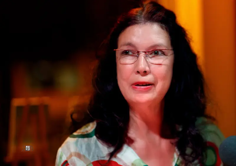 Porträtfoto von Sylvia Krupicka, sie schaut an der Kamera vorbei, hat lange, dunkle Haare und trägt eine zarte Brille, im Hintergrund ist ein Raum, draussen ist es dunkel.