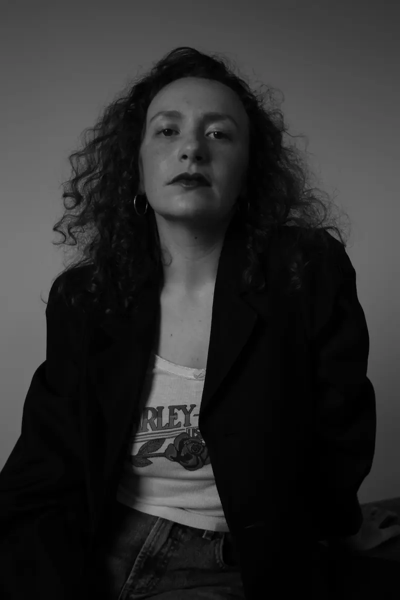 Schwarzweiß Porträtfoto von Roxie Perkins, sie hat lange, gewellte Haare, blickt leicht von oben in die Kamera hinein, sie trägt ein weißes Tshit mit einer Rose darauf und ein schwarzes Jackett