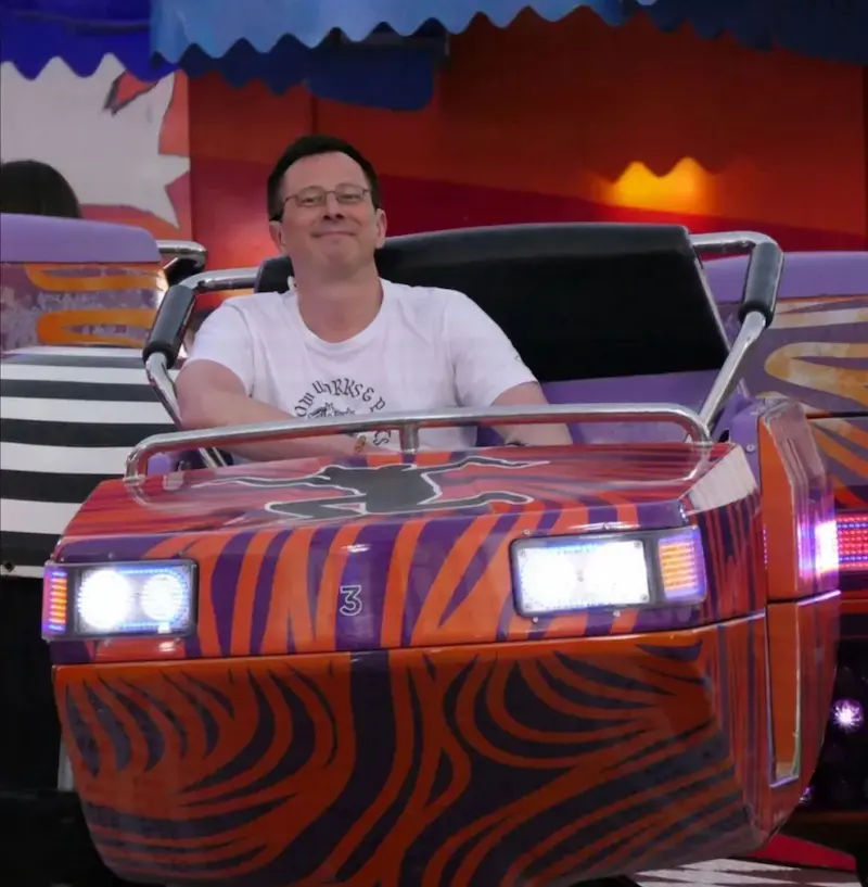 Mathias Traxler sitzt in einem Jahrmarkt Fahrgeschäft, trägt ein weißes Tshirt und lächelt in die Kamera.