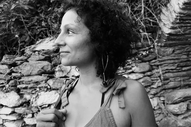 Schwarzweiß Porträtfoto von Maria Constanti. Sie schaut lächelnd zur Seite, hinter ihr ist eine überwachsene Steinmauer.