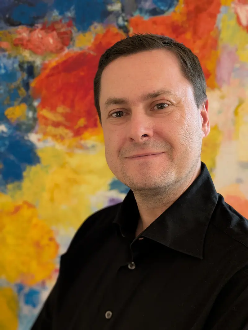 Porträtfoto von Tim Slagman, er lächelt in die Kamera,  trägt ein schwarzes Hemd, im Hintergrund ist eine bunt bemalte Leinwand.