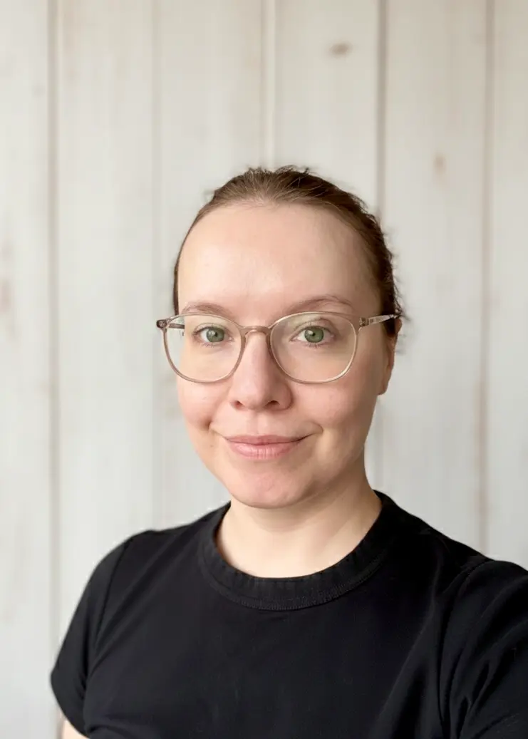 Porträtfoto von Kerstin Hörig, sie lächelt in die Kamera, trägt ein schwarzes Tshirt und eine Brille, im Hintergrund ist eine Wand mit weiß bemalten Holzlatten