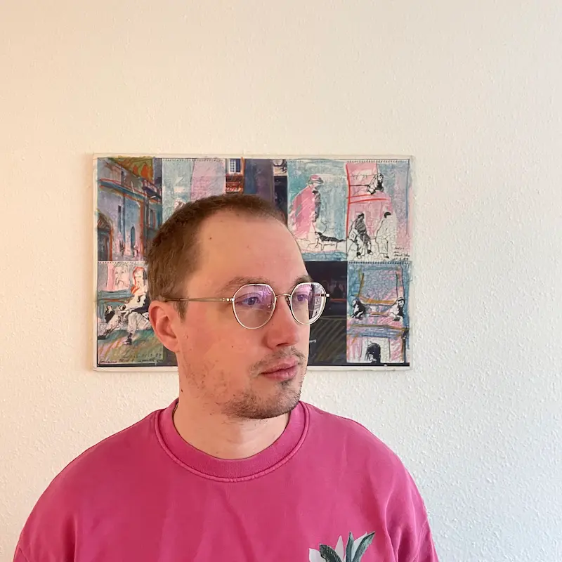  Porträtfoto von Anton Borlinghaus, er steht vor einer bunten, comichaften Zeichnung, er schaut zur Seite, trägt ein pinkes Tshirt und eine Brille.