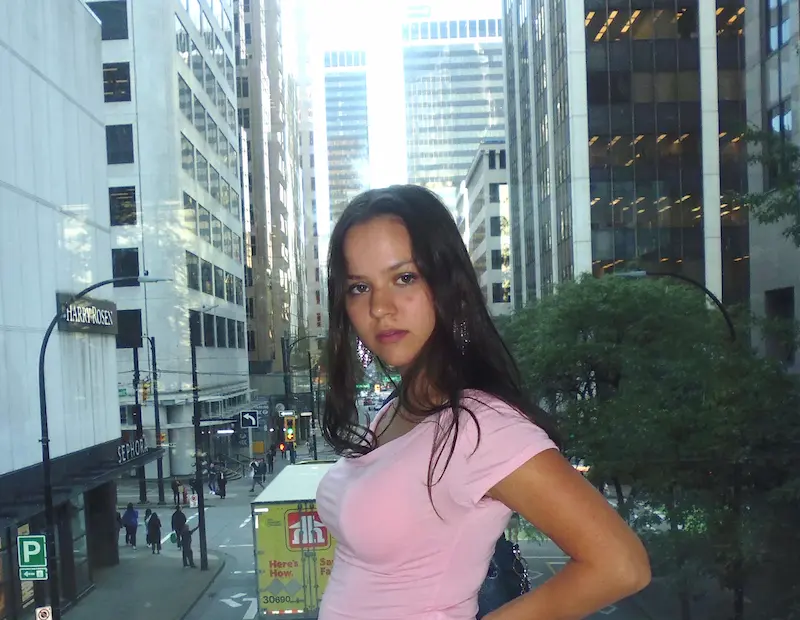 Porträtfoto von Coco Putz, sie steht seitlich zur Kamera, hat den Arm in die Hüfte gestemmt, trägt ein rosarotes Tshirt, im Hintergrund Hochhäuser