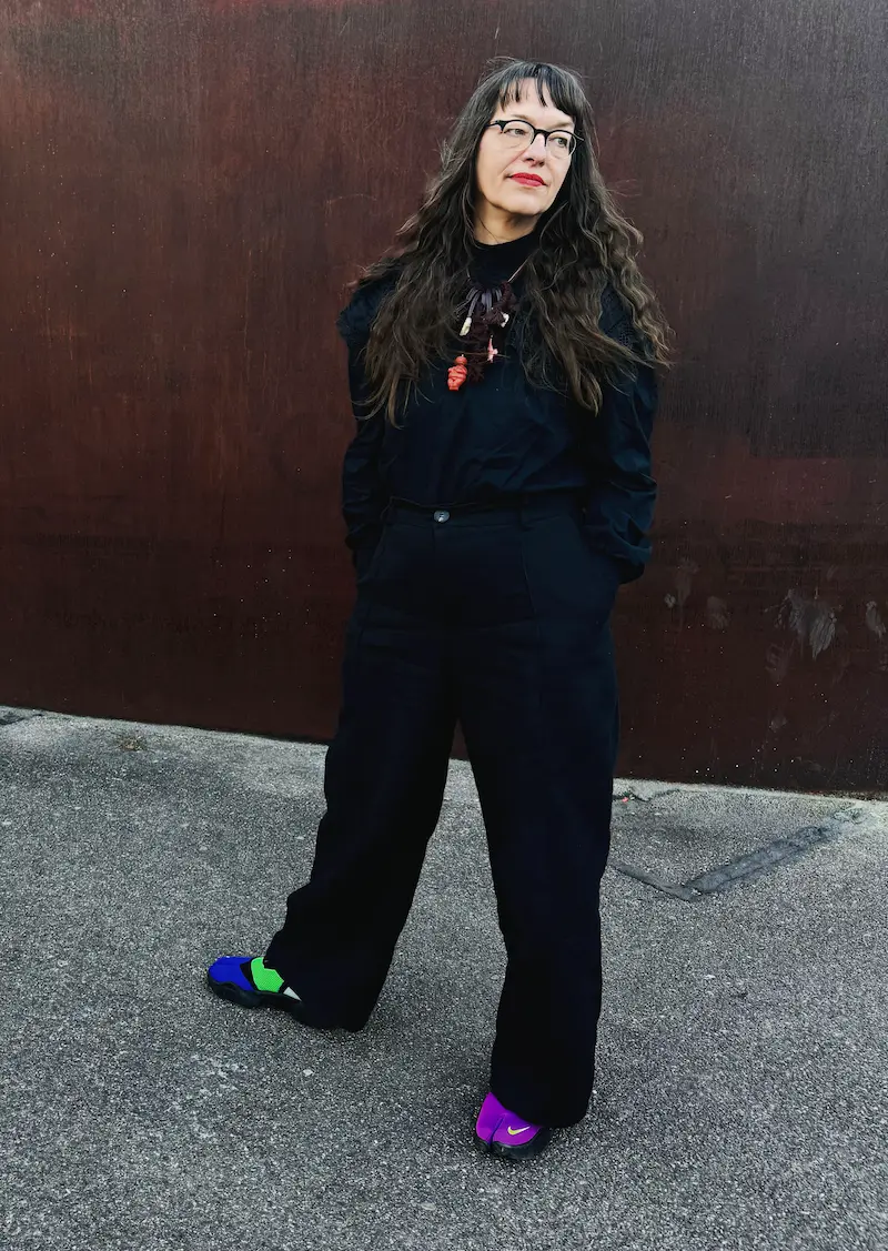 Porträtfoto von Ulli Lust, sie trägt dunkle Kleider, knallig bunte Schuhe, eine Kette, an der eine orange Figur eines nackten Frauenkörper hängt. Ihre Hände sind in ihren Hosentaschen und sie schaut lässig zur Seite.