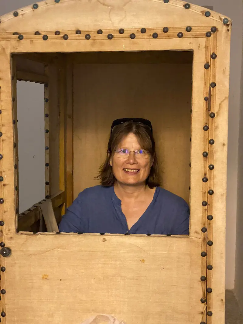 Porträtfoto von Carola Köhler, sie ist in einem kleinen Holzhäuschen und schaut lächelnd aus dem Fenster heraus.