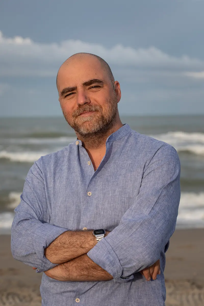 Porträtfoto von Dario Ferrari, er hat die Arme verschränkt, blickt mit einem leichten Lächeln und zusammengekniffenen Augen in die Kamera. Er trägt ein hellblaues Hemd, im Hintergrund sind ein Strand und Wellen des Meeres.
