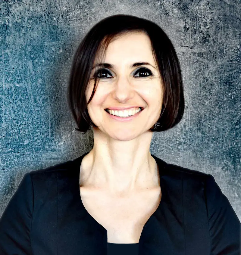 Porträtfoto von Katia Tangian. Vor einem bläulichen Hintergrund lächelt sie direkt in die Kamera. Ihre Augen sind dunkel geschminkt und sie trägt einen dunkelblauen Blazer.