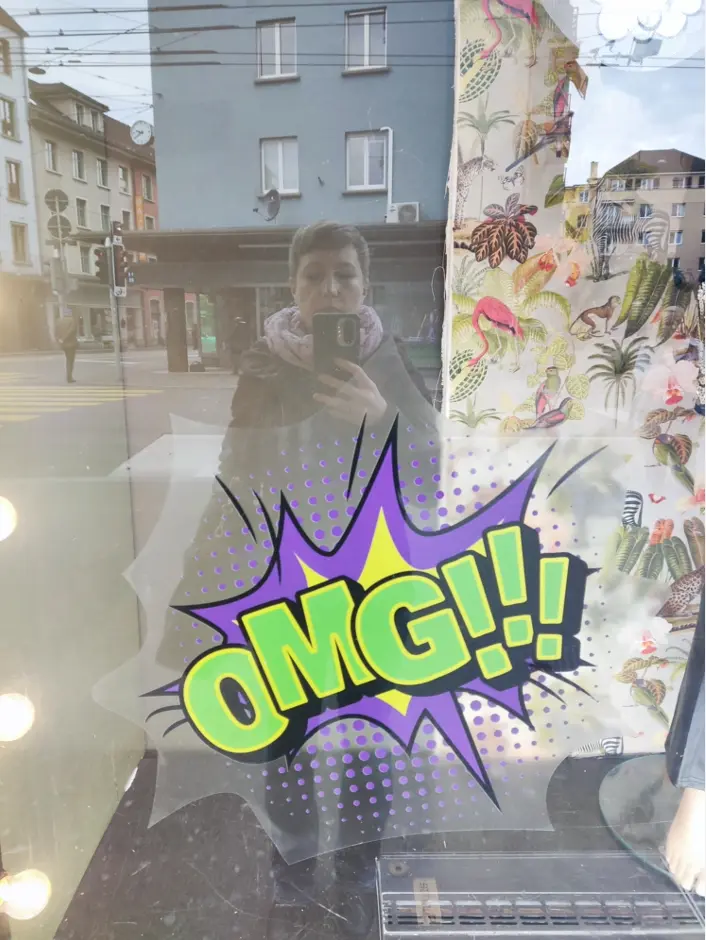 Selfie von Christiane Quandt. Sie fotografiert ihr eigenes Spiegelbild in einem Schaufenster, auf dem in bunten großen comichaften Buchstaben "OMG!!!" steht. Ihr Spiegelbild ist in blaßen Farben hinter der bunten Aufschrift zu sehen.