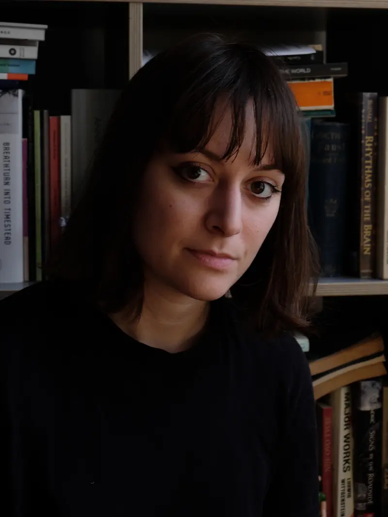 Porträtfoto von Katarina Gotic Damiani. Sie blickt ernst in die Kamera, hinter ihr ist ein Büchergestell mit wild gestapelten Büchern.