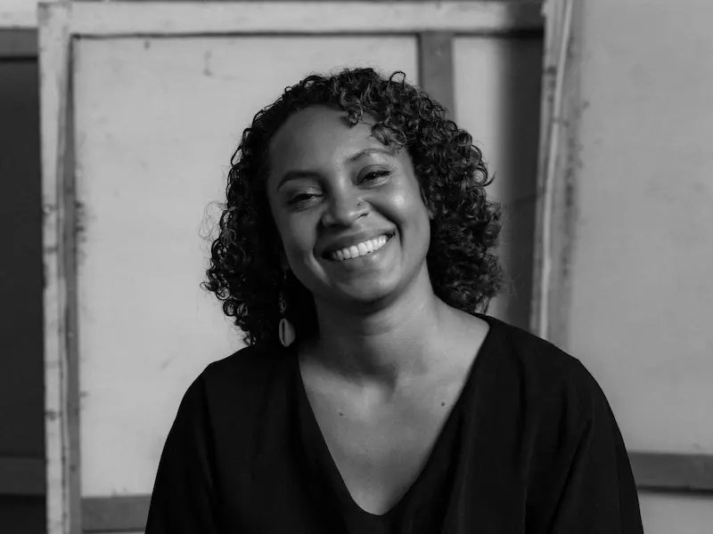 Schwarzweiß Porträtfoto von Melody Makeda Ledwon. Sie lächelt in die Kamera und steht vor einer Betonwand.