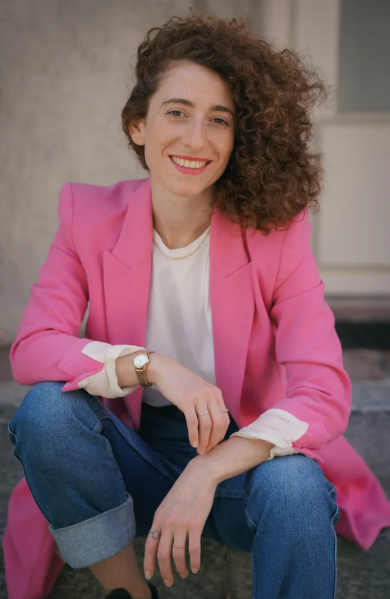 Porträtfoto von Giuliana Kiersz. Sie sitzt auf einer Treppenstufe, lächelt in die Kamera und trägt blaue Jeans und einen pinken Blazer.