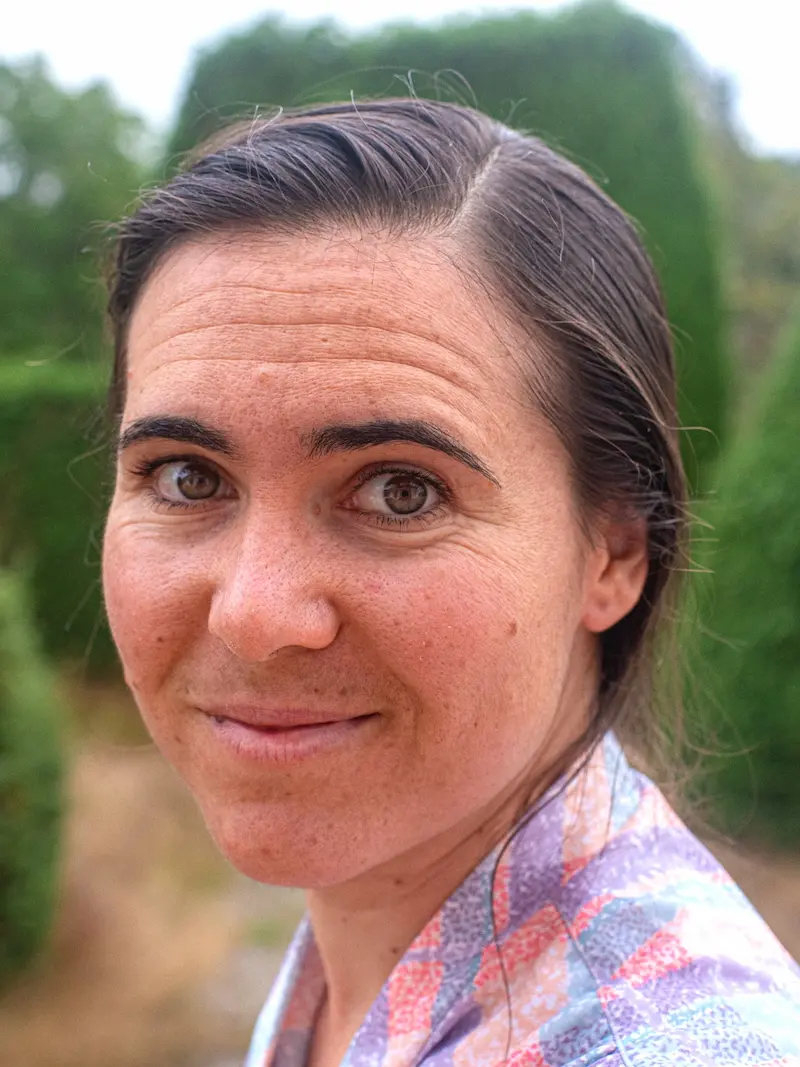 Porträtfoto von Elsa Cailletaud. Sie lächelt in die Kamera, ihre dunklen Haare sind zusammengebunden und sie trägt ein buntes Hemd.