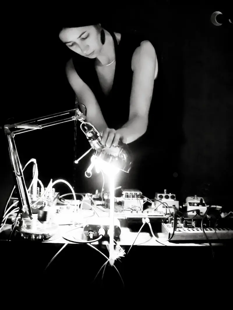 Schwarzweiß Foto von Cate Hops. Sie steht an einem Mischpult. Eine Schreibtischlampe projiziert einen hellen Lichtstrahl auf den Tisch, auf dem zahlreiche Geräte mit vielen Schaltern und Knöpfen stehen.