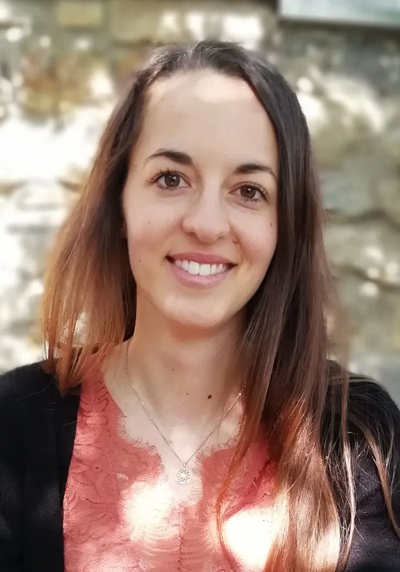 Porträtfoto von Annika Posselt. Sie lächelt in die Kamera, der Hintergrund ist verschwommen, die Sonne scheint. Sie trägt ihre langen, braunen Haar offen und sie trägt eine rosafarbene Bluse mit einer schwarzen Strickjacke.