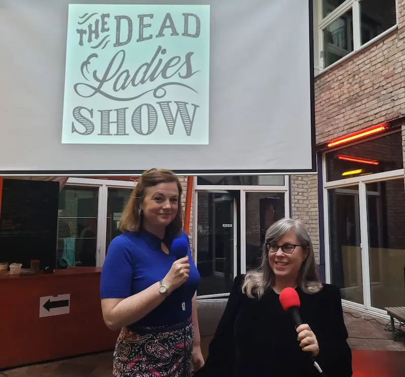 Foto von Katy Derbyshire und Susan Stone. Sie stehen vor einer Leinwand, auf der das Logo der Dead Ladies Show projiziert ist. Sie halten beide Mikrophone in den Händen und lächeln in diea Kamera.
