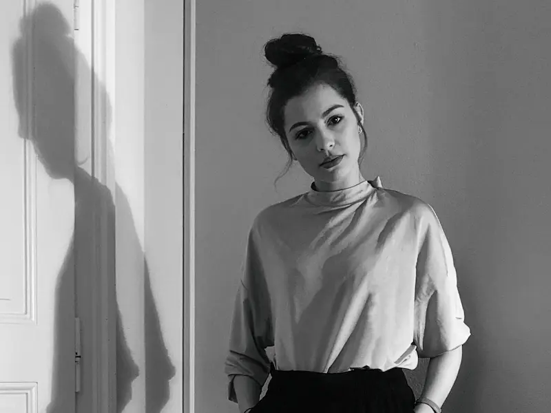 Schwarzweiß Foto von Lea Kubeneck. Sie schaut ernst in die Kamera und trägt ihre dunklen Haare in einem Dutt zusammengebunden. Neben ihr ist eine weiße Türe, uf die ihr Schatten fällt.