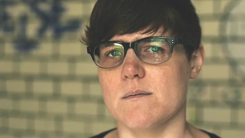 Porträtfoto von Kate McNaughton. Sie steht vor einer Mauer, die mit Graffiti bemalt ist. Sie blickt ernst in die Kamera, trägt eine dunkle Brille mit einem dicken Rahmen und kurze schwarze Haare.