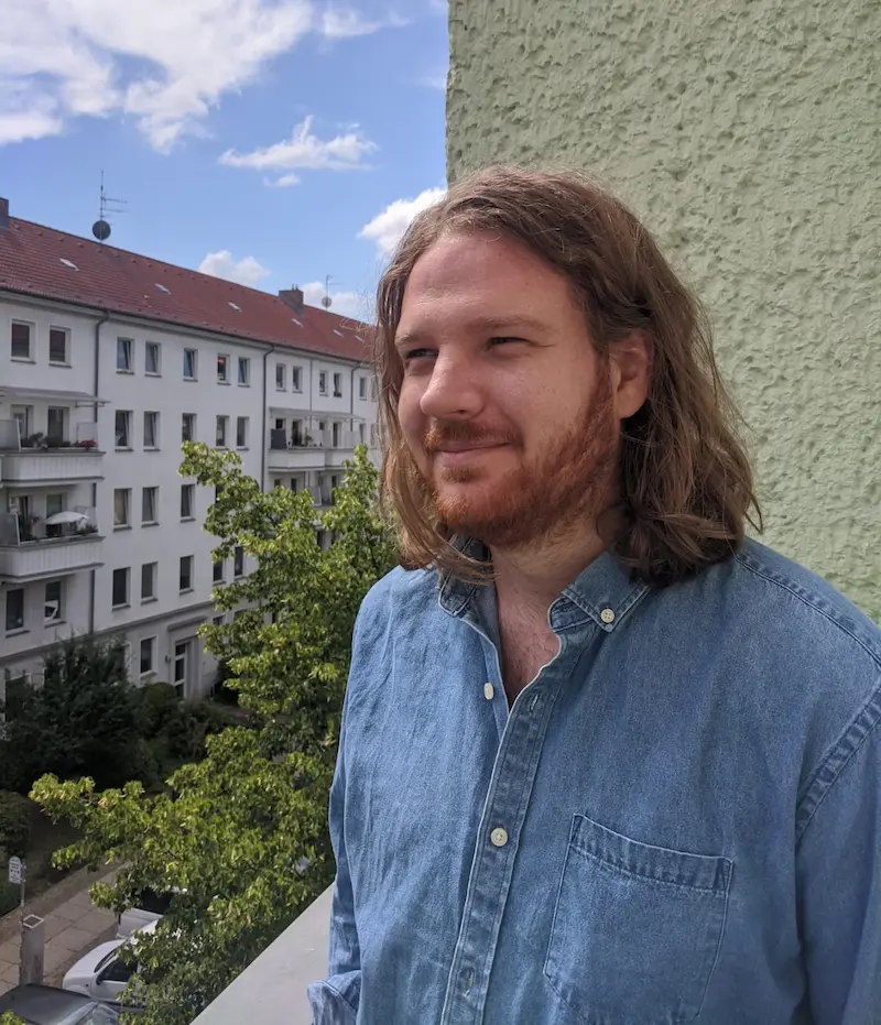 Foto von Alex Wells. Er steht auf einem Balkon und blickt in die Ferne. Das Wetter ist schön, Alex Wells lächelt. Er trägt ein Jeanshemd und seine rötlichen Haare hängen ihm über die Schultern.