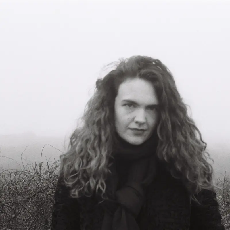 Ein Porträtfoto von Madeleine Watts. Die Autorin sieht in die Kamera, lange Locken fallen über ihre Schultern, der Kopf leicht zur Seite geneigt. In schwarz-weiß, im Hintergrund karge Büsche, winterliche Stimmung.