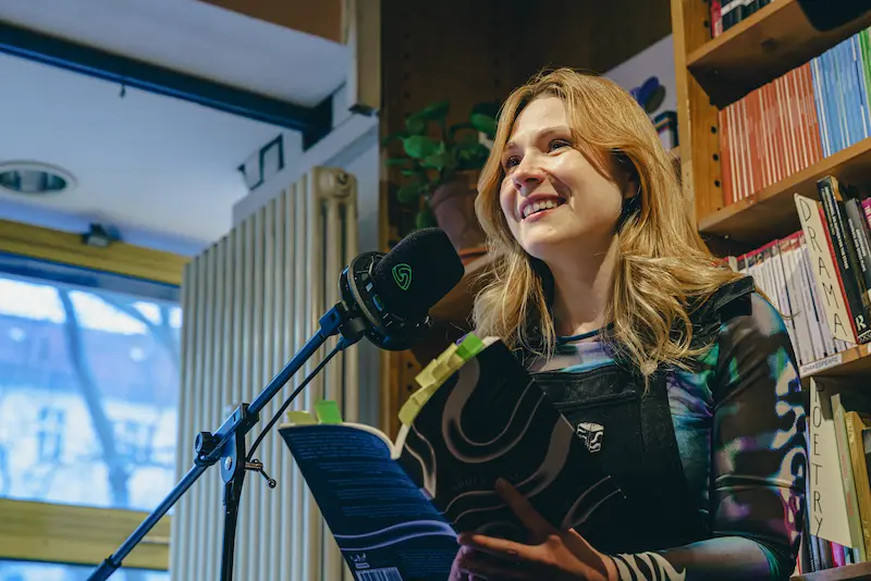 Demi Anter liest aus einem Buch, hinter ihr ein Bücherregal. Die langen blonden Haare liegen auf ihrem bunten Shirt, sie spricht in das Mikrofon und lächelt in die Menge.