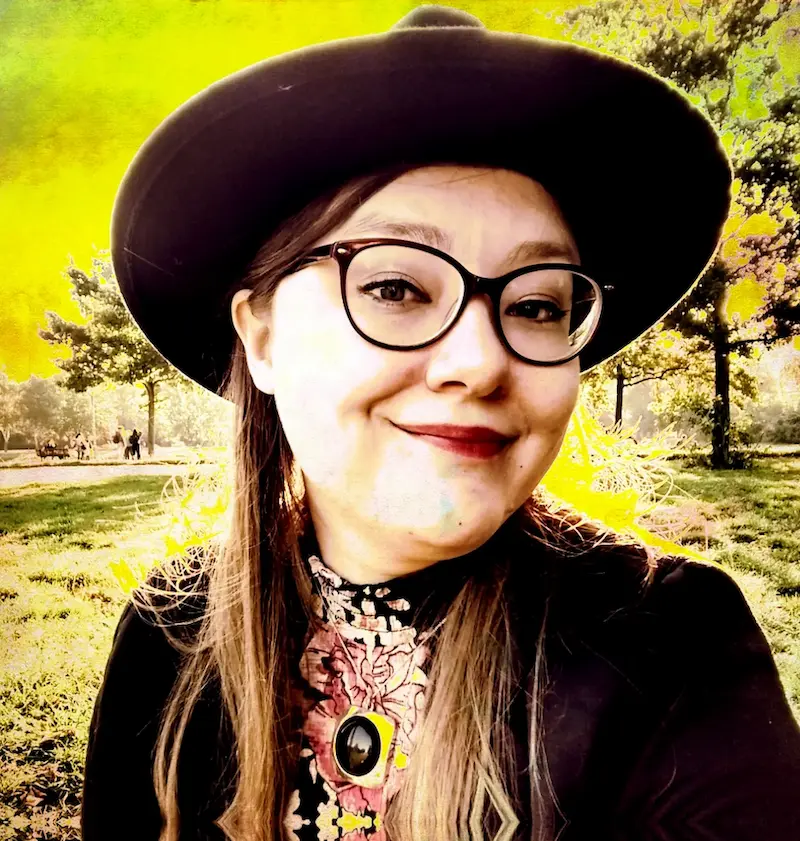 Riikka Korpela trägt einen großen, dunklen Hut und eine dunkle Brille. Die Landschaft im Hintergrund ist gelb gefärbt, sie dreht sich leicht lächelnd zur Seite.
