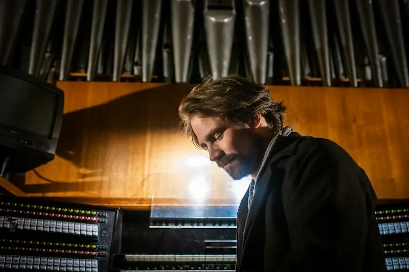 Kit Downes, umgeben von Instrumenten, blickt im schwarzen Anzug nach unten, der Körper zur Seite geneigt.