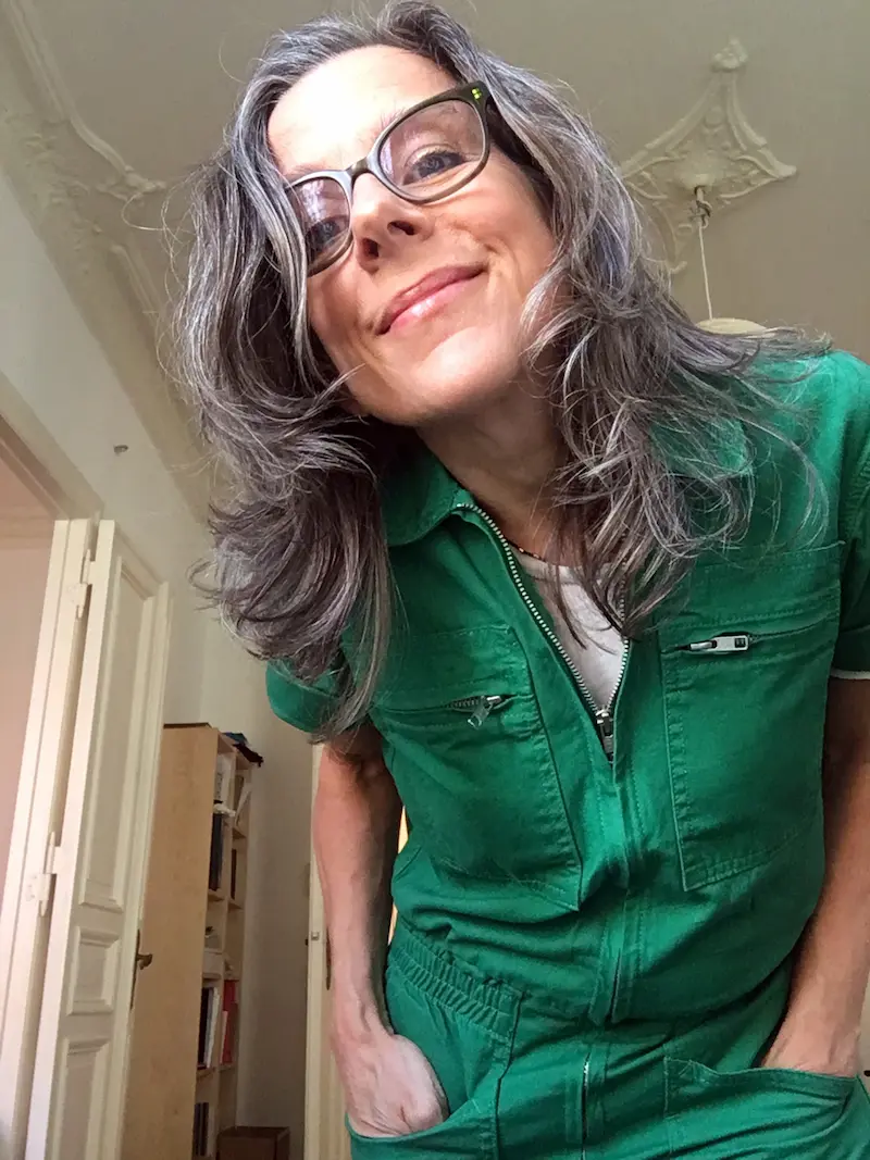 Nina Katchadourian beugt sich zur Kamera, die Hände lässig in den grünen Overall gesteckt. Ihre dunklen Haar hängen nach vorne, sie lächelt, trägt eine dunkle Brille.