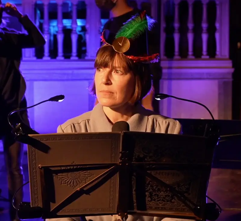 Sally O'Reilly sitzt an einem Instrument, vermutlich ein Piano, vor ihr der Notenständer. Sie trägt einen kleinen Hut mit Feder, einen Pony, blickt zur Seite.