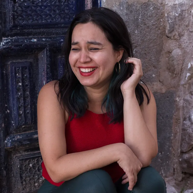 Ein Porträt von Susana Vásquez Torres. Sie blickt zur Seite, lacht, dunkle Haare. Roter Lippenstift und rotes Top.