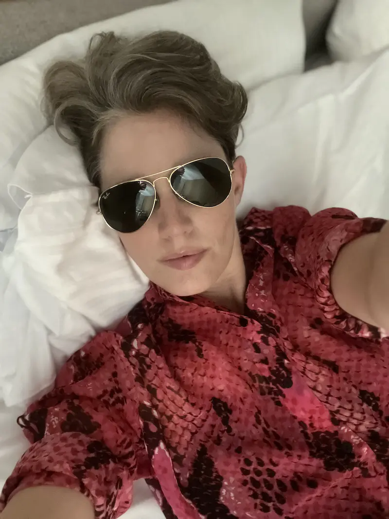 Ein Selfie von Jana Petersen. Sie trägt eine Sonnenbrille, rotes Hemd, liegt auf einem Bett.