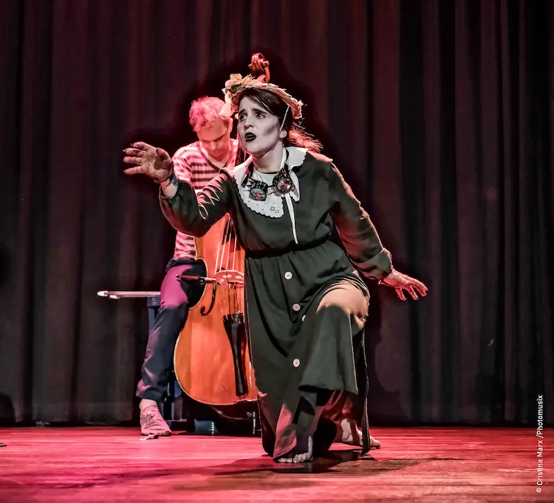 Künstlerin in grünem Kleid und mit Hut performed auf Theaterbühne, hinter ihr ein Cellist