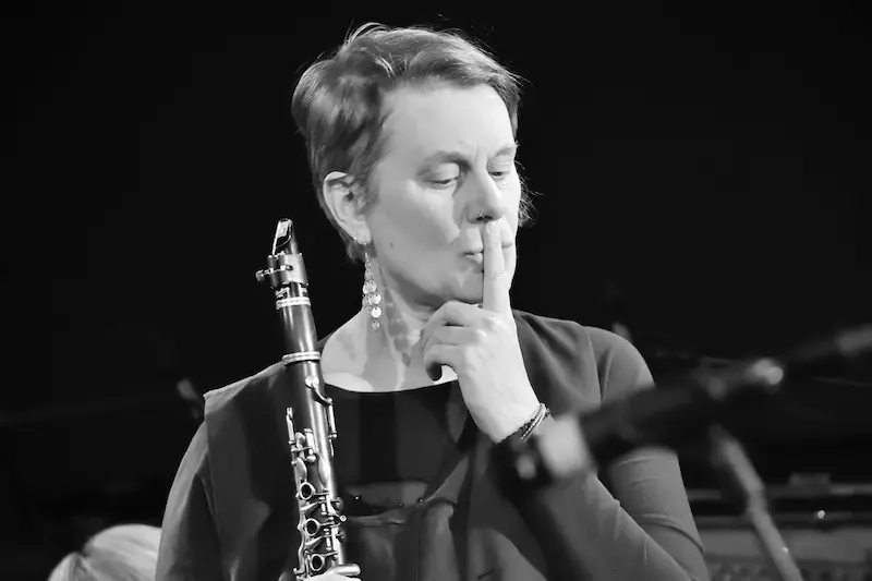 schwarz-weiß Bild der Künstlerin, hält in einer Hand eine Klarinette und den Zeigfinger der anderen an den Mund