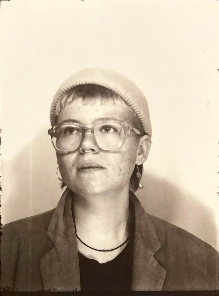 Schwarz-weiß-Foto, Laa Wa ha kurze helle Haare und trägt eine Brille mit großen Gläsern, einehelle Mütze und ein dunkles Jacket.