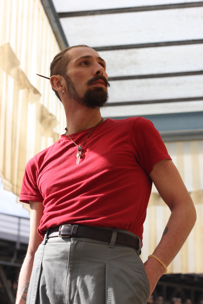Eyk Kauly hat dunkle Haare und trägt einen gepflegten Schnurrbart und Kinnbart. Er hat eine graue Hose und ein rotes T-Shirt an und trägt eine Kette mit langem Anhänger.