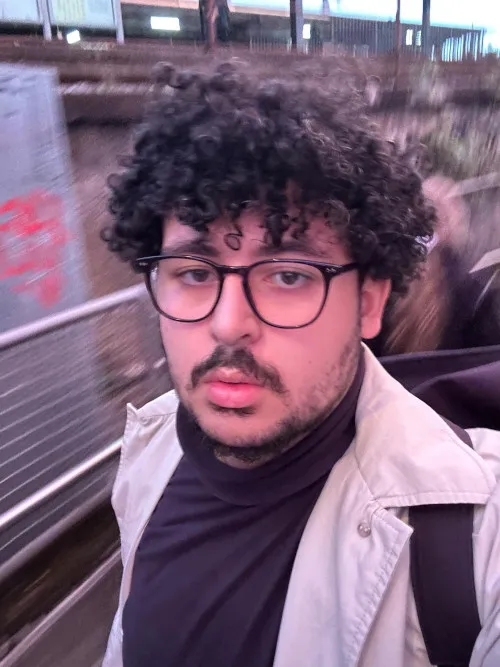 Sedat Ebubekir Günes hat schwarze Locken und trägt eine Brille mit dunklem Gestell, ein schwarzes Rollkragenshirt und eine weiße Jacke.