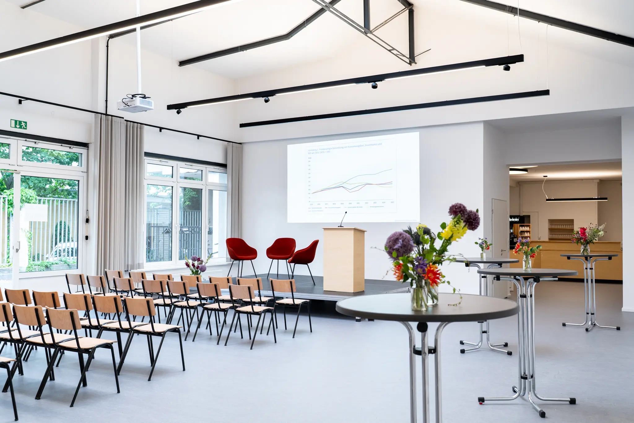 Bühne mit Stehpult und drei roten Sesseln, davor leere Stuhlreihen und rechts davon runde Stehtische mit Blumensträußen