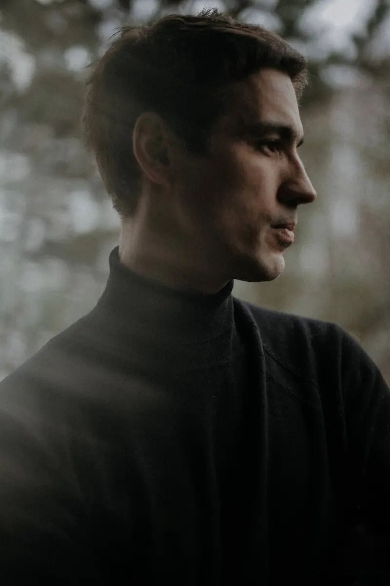 Portrait of the artist wearing black turtle neck, looking to the side, trees in the background