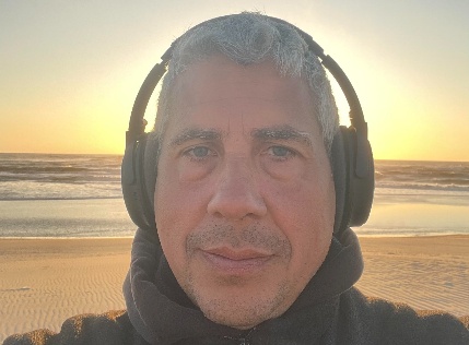 Tobias Breyer trägt Kopfhörer und steht vor einem Sonnenuntergang am Strand.