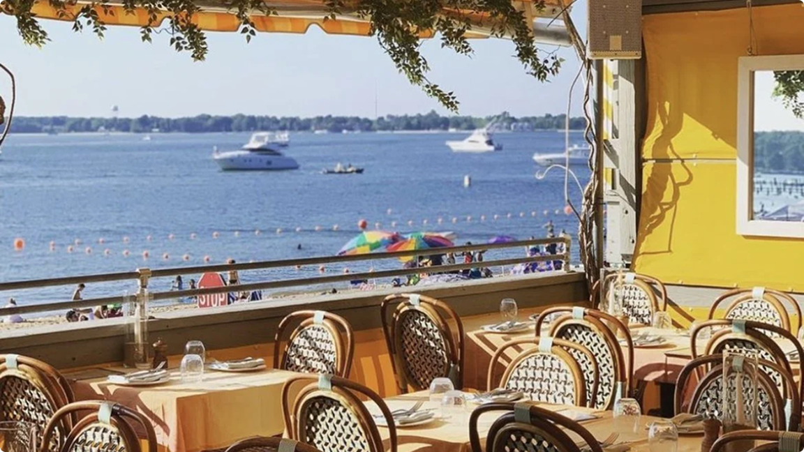Outdoor restaurant seating with wooden chairs and tables overlooking a beach with colorful umbrellas and boats on the water.