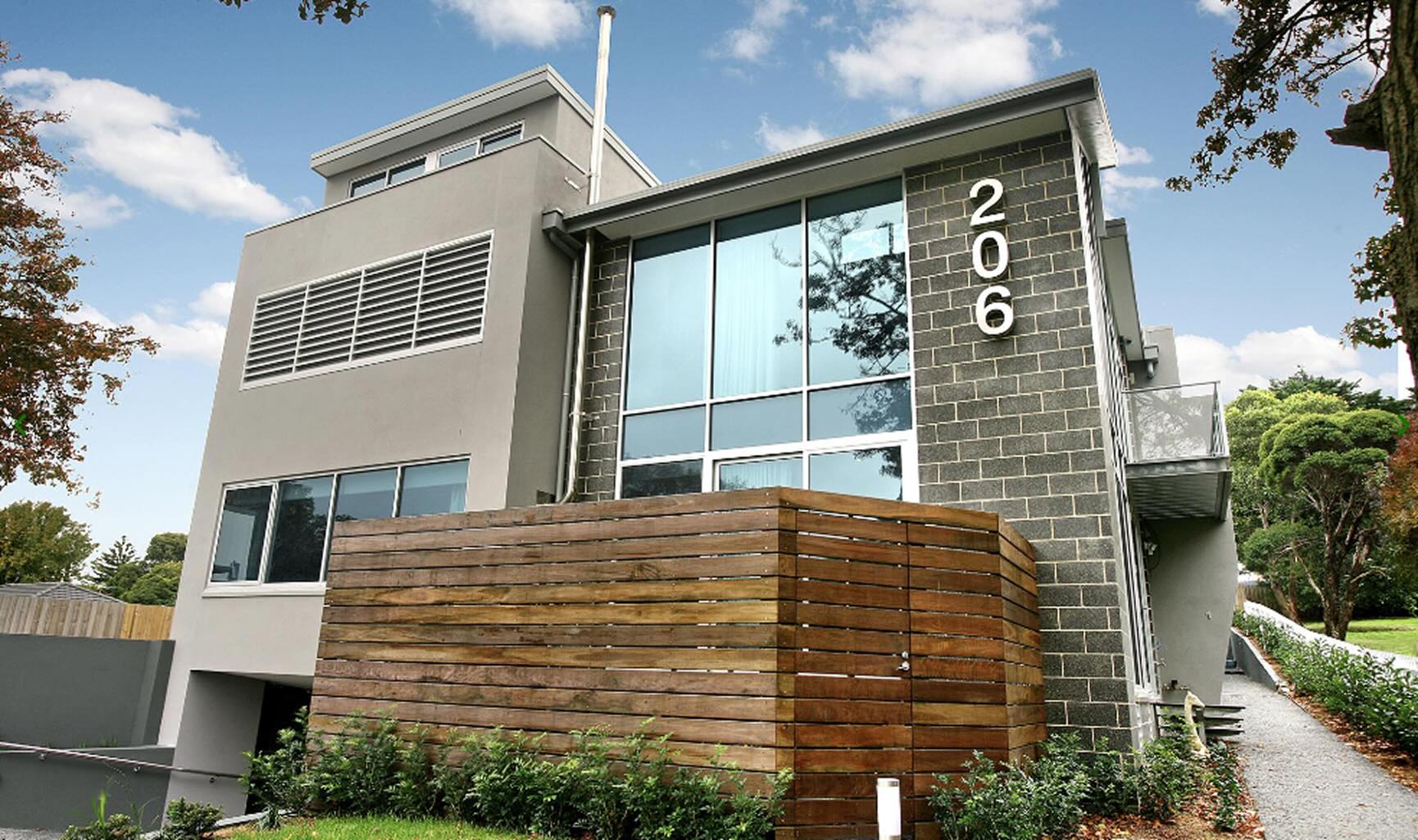 Modern two-story house with large windows, gray brick and stucco walls, wood privacy fence, and the number 206 displayed vertically.