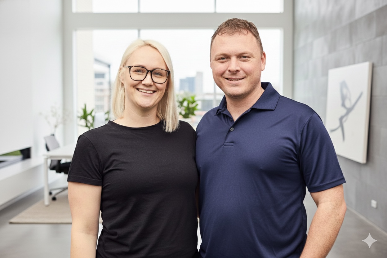 Smiling man in a navy blue polo shirt and woman wearing glasses and a black t-shirt standing close together in a bright modern office.