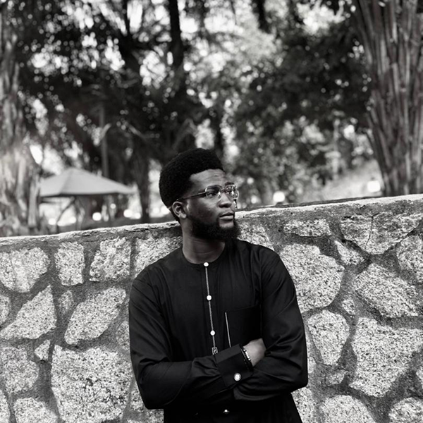 A black-and-white portrait of Abdur-Rasheed with glasses and a beard, wearing a traditional dark outfit, standing with his arms crossed in front of a stone wall and trees in the background.