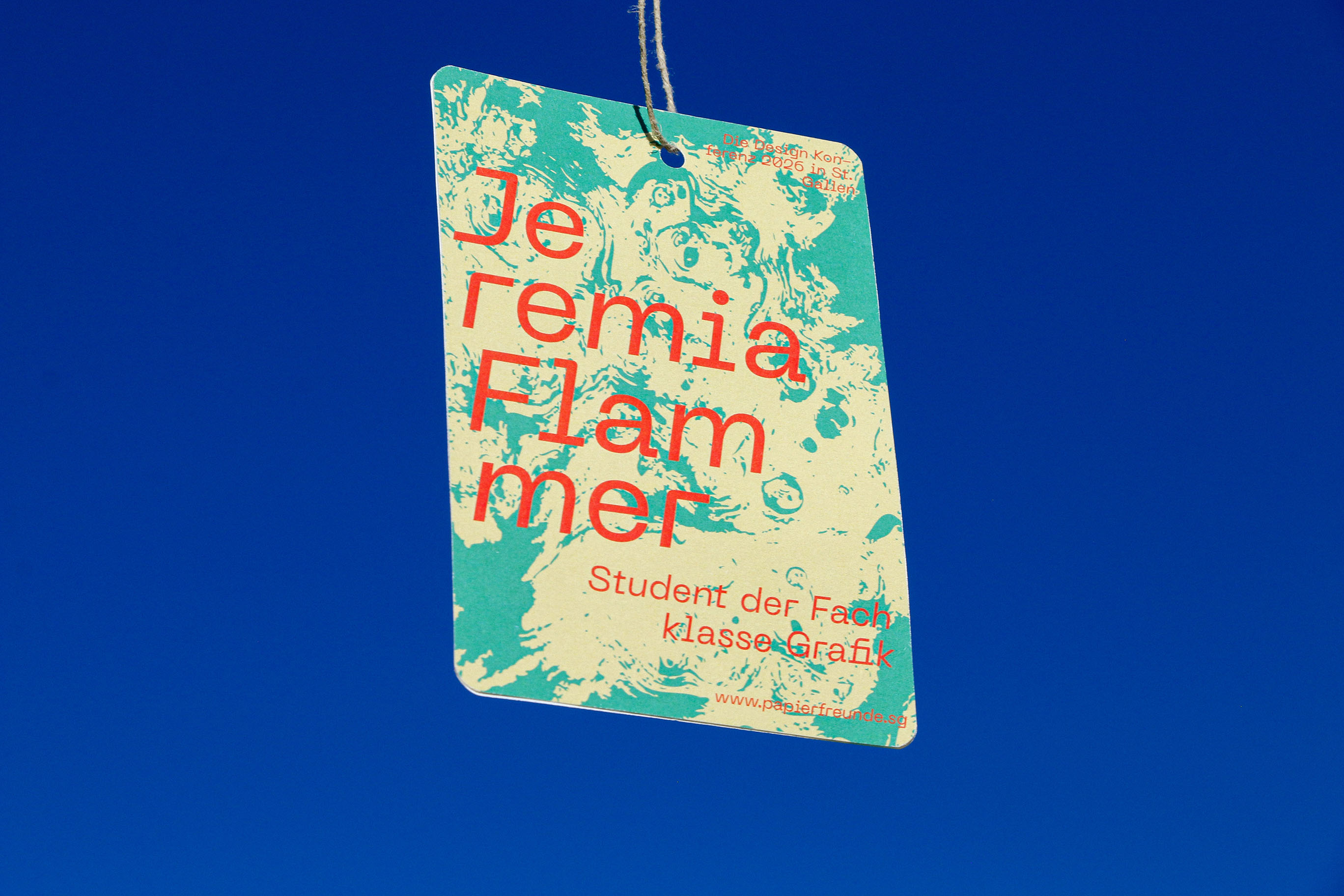 Hanging tag with turquoise and beige marbled background reading 'Jeremia Flammer' and 'Student der Fachklasse Grafik' against a clear blue sky.